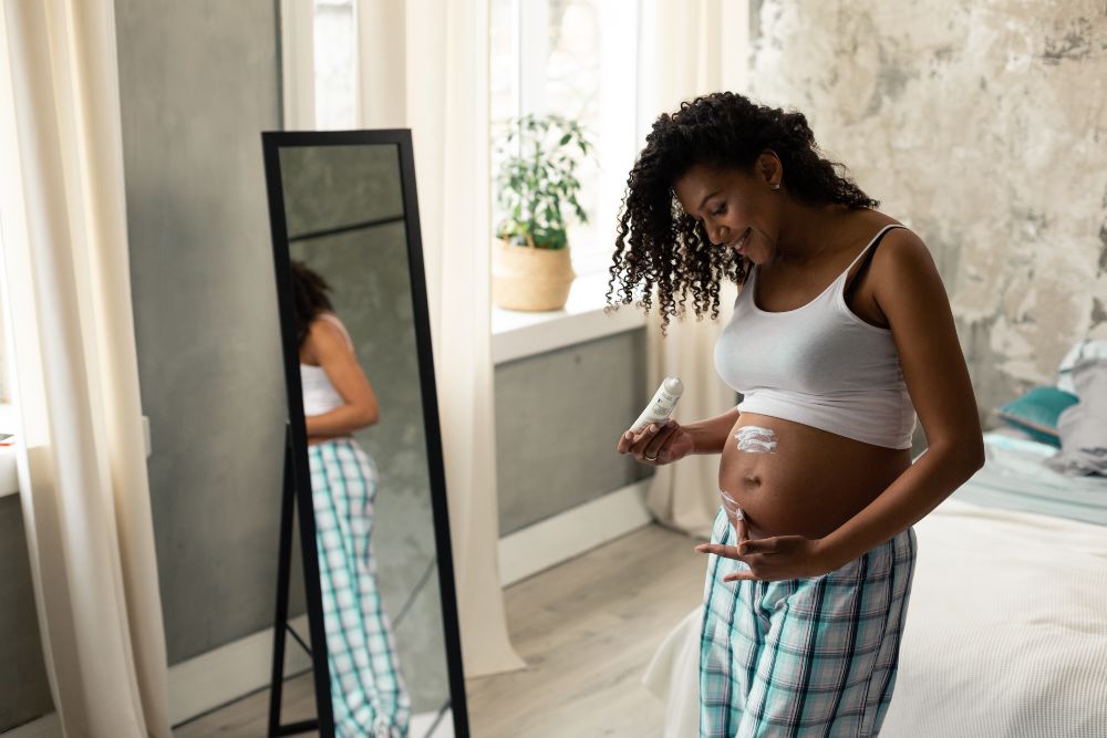 Mulher passando creme na barriga de grávida em frente ao espelho buscando autoestima feminina